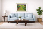 Oak framed wall art hanging in a lLiving room with a light blue sofa in a living room with a painting, a table, and plant.